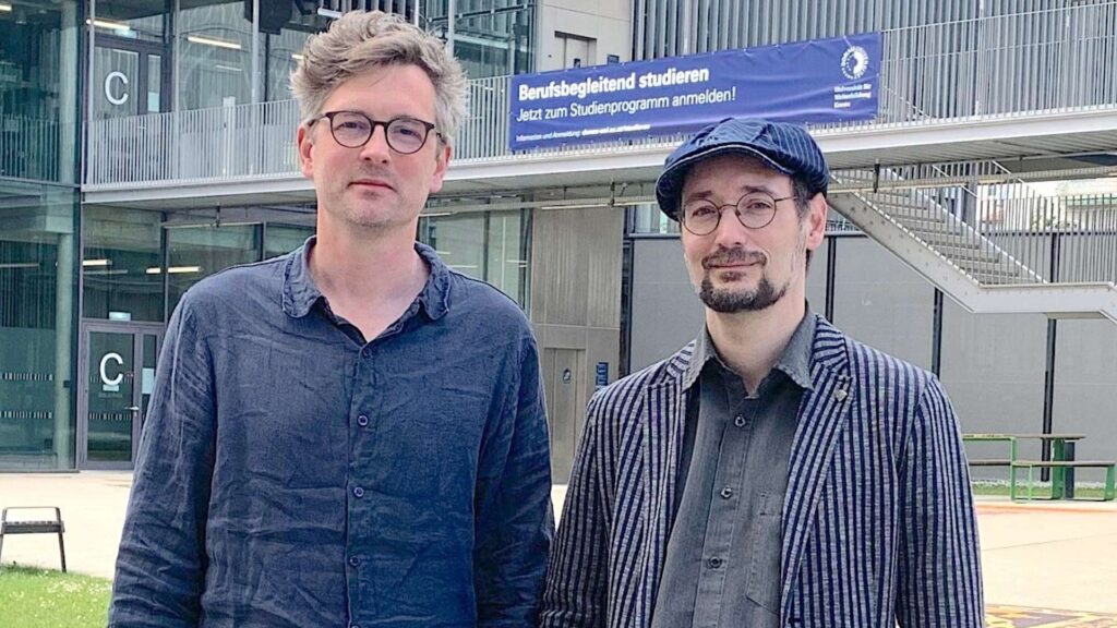 Two men pose outside a modern glass building; one wears a blue shirt, the other a striped blazer and cap, both with glasses.